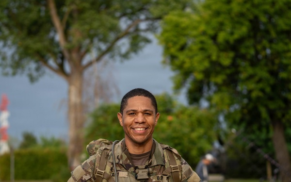 A smiling U.S. Army soldier in MultiCam/OCP camouflage uniform is posing outdoors while holding a clear plastic bag containing what appears to be military patches or insignia. He is wearing a rucksack and standing on a brick pathway near a building, trees, and red-and-white bollards. The setting appears to be a military installation or base, likely in Europe.
