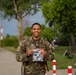 A smiling U.S. Army soldier in MultiCam/OCP camouflage uniform is posing outdoors while holding a clear plastic bag containing what appears to be military patches or insignia. He is wearing a rucksack and standing on a brick pathway near a building, trees, and red-and-white bollards. The setting appears to be a military installation or base, likely in Europe.
