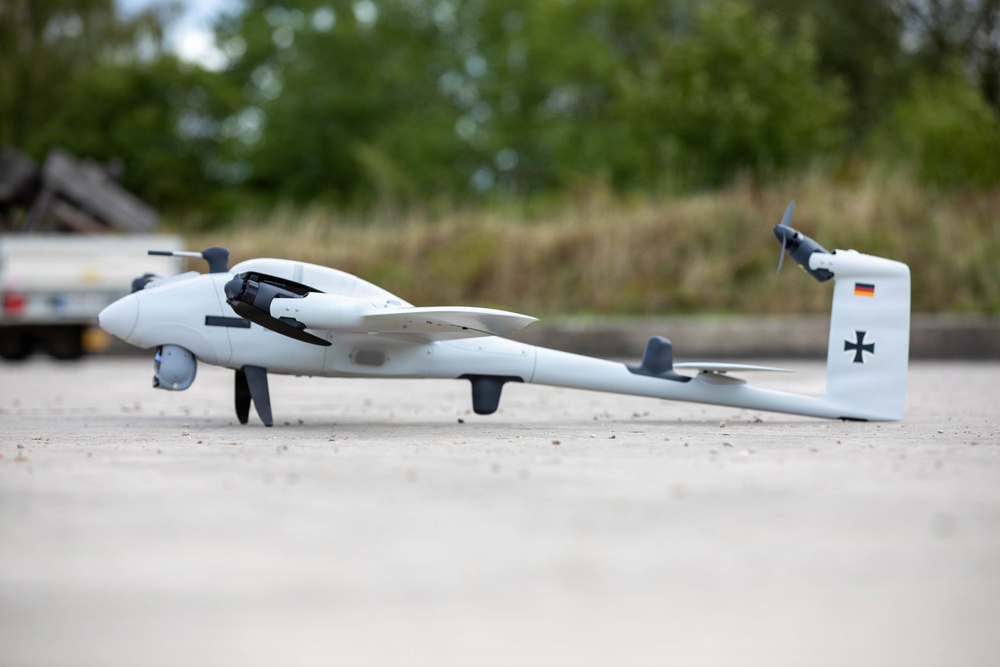 The image shows a small fixed-wing UAV (unmanned aerial vehicle) or drone resting on a paved surface. The aircraft bears German military markings, including the Bundeswehr Iron Cross and the German flag on its tail fin. It appears to be a VTOL (Vertical Take-Off and Landing) capable drone with multiple rotors and a pusher propeller configuration.
