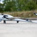 The image shows a small fixed-wing UAV (unmanned aerial vehicle) or drone resting on a paved surface. The aircraft bears German military markings, including the Bundeswehr Iron Cross and the German flag on its tail fin. It appears to be a VTOL (Vertical Take-Off and Landing) capable drone with multiple rotors and a pusher propeller configuration.