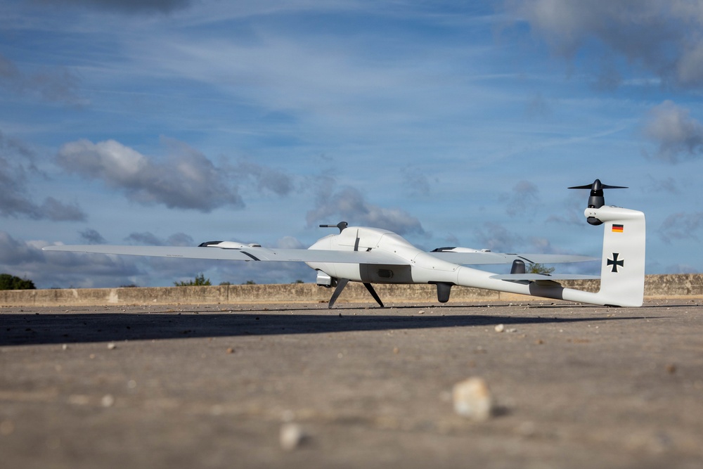 A white unmanned aerial vehicle (UAV/drone) is parked on a concrete surface outdoors. The aircraft displays a German flag and an Iron Cross (Bundeswehr insignia) on its tail fin, identifying it as a German military drone. The pusher-propeller configuration and twin-boom design are clearly visible against a partly cloudy sky.
