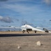 A white unmanned aerial vehicle (UAV/drone) is parked on a concrete surface outdoors. The aircraft displays a German flag and an Iron Cross (Bundeswehr insignia) on its tail fin, identifying it as a German military drone. The pusher-propeller configuration and twin-boom design are clearly visible against a partly cloudy sky.