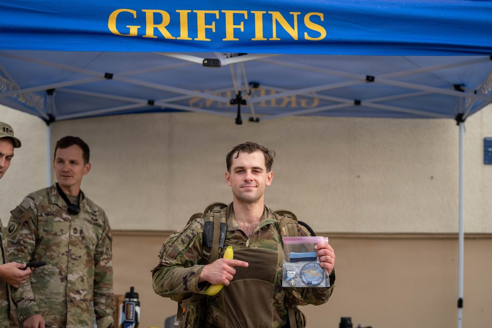 A military service member stands under a blue 'GRIFFINS' branded canopy tent, holding a banana and a clear plastic bag containing what appears to be a patch or coin and other small items. He is wearing camouflage tactical gear with a plate carrier and appears to have been physically active, evidenced by his disheveled appearance. Two other uniformed personnel are visible in the background.