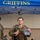 12th Combat Aviation Brigade hosts Danish Flyver March A military service member stands under a blue 'GRIFFINS' branded canopy tent, holding a banana and a clear plastic bag containing what appears to be a patch or coin and other small items. He is wearing camouflage tactical gear with a plate carrier and appears to have been physically active, evidenced by his disheveled appearance. Two other uniformed personnel are visible in the background.