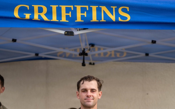 A military service member stands under a blue 'GRIFFINS' branded canopy tent, holding a banana and a clear plastic bag containing what appears to be a patch or coin and other small items. He is wearing camouflage tactical gear with a plate carrier and appears to have been physically active, evidenced by his disheveled appearance. Two other uniformed personnel are visible in the background.
