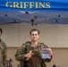 A military service member stands under a blue 'GRIFFINS' branded canopy tent, holding a banana and a clear plastic bag containing what appears to be a patch or coin and other small items. He is wearing camouflage tactical gear with a plate carrier and appears to have been physically active, evidenced by his disheveled appearance. Two other uniformed personnel are visible in the background.