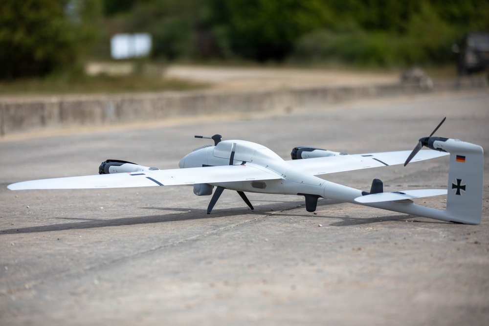 A white fixed-wing UAV (unmanned aerial vehicle) with VTOL (vertical take-off and landing) rotors is positioned on a paved surface outdoors. The aircraft displays German military markings, including the Bundeswehr Iron Cross and the German flag on its tail fin. The drone appears to be in a pre-flight or post-flight ground state.