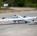 A white fixed-wing UAV (unmanned aerial vehicle) with VTOL (vertical take-off and landing) rotors is positioned on a paved surface outdoors. The aircraft displays German military markings, including the Bundeswehr Iron Cross and the German flag on its tail fin. The drone appears to be in a pre-flight or post-flight ground state.