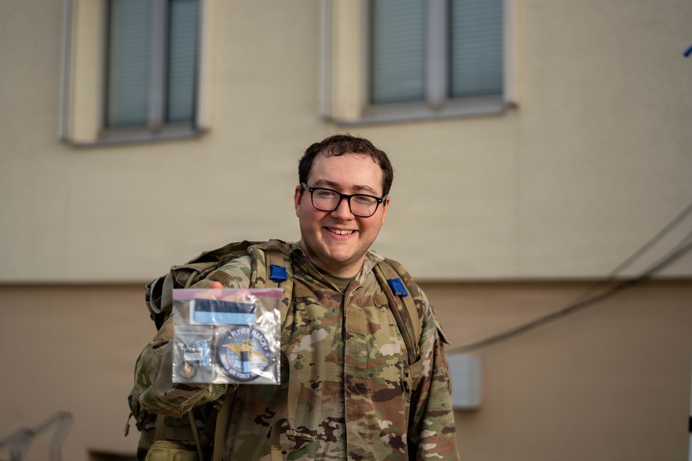 A smiling military service member in OCP (Operational Camouflage Pattern) uniform is holding up a clear plastic bag containing what appears to be a challenge coin or patch related to a 'Flyer March' Air Force event. They are wearing glasses and carrying a tactical backpack with blue rank or unit insignia patches visible. The photo appears to be taken outdoors in front of a light-colored building.