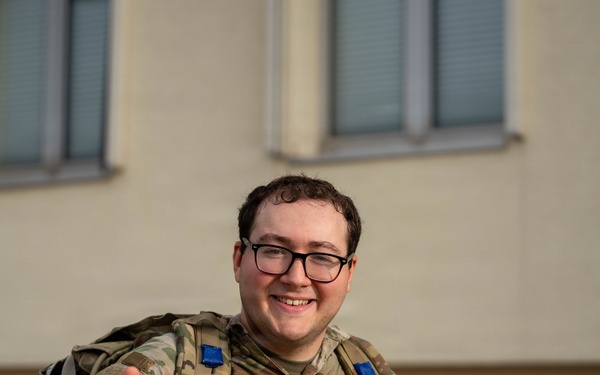 A smiling military service member in OCP (Operational Camouflage Pattern) uniform is holding up a clear plastic bag containing what appears to be a challenge coin or patch related to a 'Flyer March' Air Force event. They are wearing glasses and carrying a tactical backpack with blue rank or unit insignia patches visible. The photo appears to be taken outdoors in front of a light-colored building.