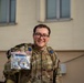 A smiling military service member in OCP (Operational Camouflage Pattern) uniform is holding up a clear plastic bag containing what appears to be a challenge coin or patch related to a 'Flyer March' Air Force event. They are wearing glasses and carrying a tactical backpack with blue rank or unit insignia patches visible. The photo appears to be taken outdoors in front of a light-colored building.