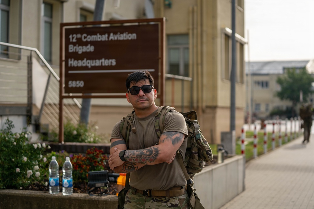 A male soldier in U.S. Army combat uniform stands with arms crossed in front of the 12th Combat Aviation Brigade Headquarters sign, numbered 5850. He is wearing sunglasses, a tactical backpack, and has visible tattoos on his right forearm. The setting appears to be a military installation, likely in Europe based on the architectural style.