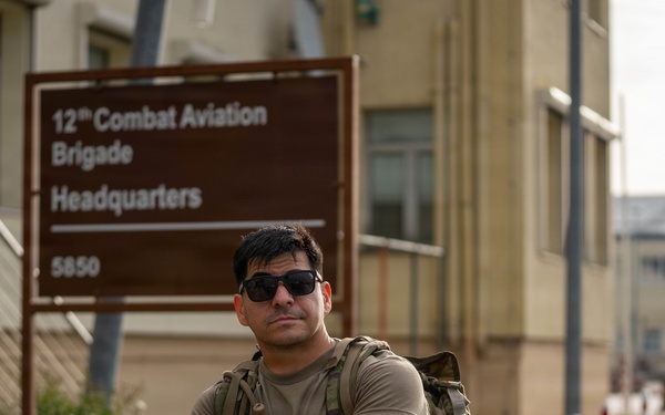 A male soldier in U.S. Army combat uniform stands with arms crossed in front of the 12th Combat Aviation Brigade Headquarters sign, numbered 5850. He is wearing sunglasses, a tactical backpack, and has visible tattoos on his right forearm. The setting appears to be a military installation, likely in Europe based on the architectural style.