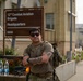 A male soldier in U.S. Army combat uniform stands with arms crossed in front of the 12th Combat Aviation Brigade Headquarters sign, numbered 5850. He is wearing sunglasses, a tactical backpack, and has visible tattoos on his right forearm. The setting appears to be a military installation, likely in Europe based on the architectural style.