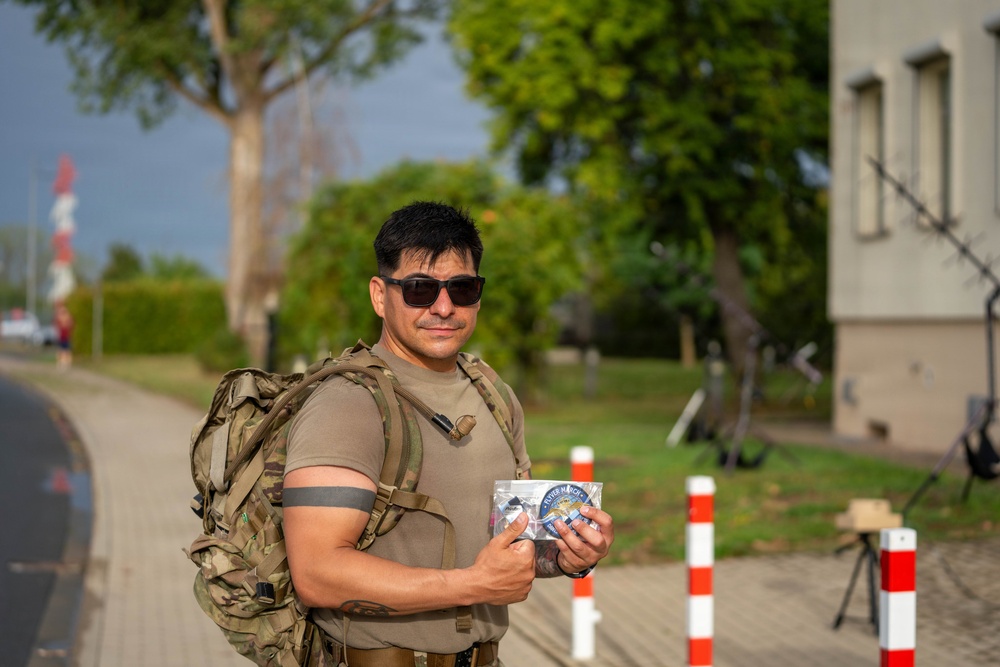 A man wearing military-style clothing and a camouflage backpack stands on a paved path, holding a small packaged food or snack item. He is wearing sunglasses and a olive/tan t-shirt, with red-and-white bollards and a building visible in the background. The setting appears to be a military base or checkpoint area with trees and green surroundings.