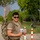 12th Combat Aviation Brigade hosts Danish Flyver March A man wearing military-style clothing and a camouflage backpack stands on a paved path, holding a small packaged food or snack item. He is wearing sunglasses and a olive/tan t-shirt, with red-and-white bollards and a building visible in the background. The setting appears to be a military base or checkpoint area with trees and green surroundings.