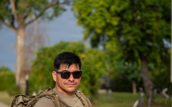 A man wearing military-style clothing and a camouflage backpack stands on a paved path, holding a small packaged food or snack item. He is wearing sunglasses and a olive/tan t-shirt, with red-and-white bollards and a building visible in the background. The setting appears to be a military base or checkpoint area with trees and green surroundings.