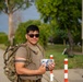 A man wearing military-style clothing and a camouflage backpack stands on a paved path, holding a small packaged food or snack item. He is wearing sunglasses and a olive/tan t-shirt, with red-and-white bollards and a building visible in the background. The setting appears to be a military base or checkpoint area with trees and green surroundings.