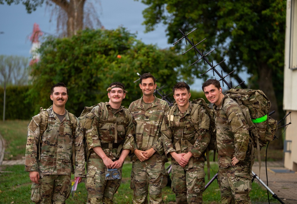 Five U.S. Army soldiers in OCP (Operational Camouflage Pattern) uniforms pose together for a group photo outdoors. The center soldier's name tape reads 'GRANT' and a 'U.S. ARMY' tape is visible. They appear to be at a military installation or training area, with a Yagi-style antenna array visible in the background.