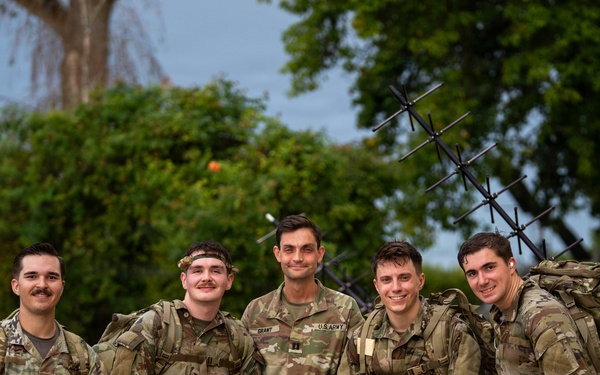 Five U.S. Army soldiers in OCP (Operational Camouflage Pattern) uniforms pose together for a group photo outdoors. The center soldier's name tape reads 'GRANT' and a 'U.S. ARMY' tape is visible. They appear to be at a military installation or training area, with a Yagi-style antenna array visible in the background.