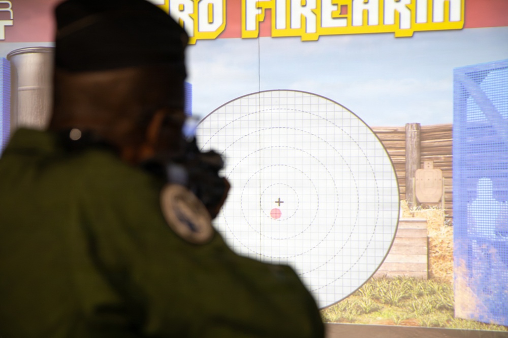 A person in military or law enforcement uniform is using a firearm simulator, aiming at a large projected screen displaying a virtual shooting range target. The screen shows a circular bullseye target with a crosshair/aiming reticle near the center, along with a simulated outdoor range environment in the background. Text at the top of the screen partially reads 'FIREARM', indicating this is a firearms training simulation system.