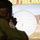 A person in military or law enforcement uniform is using a firearm simulator, aiming at a large projected screen displaying a virtual shooting range target. The screen shows a circular bullseye target with a crosshair/aiming reticle near the center, along with a simulated outdoor range environment in the background. Text at the top of the screen partially reads 'FIREARM', indicating this is a firearms training simulation system.