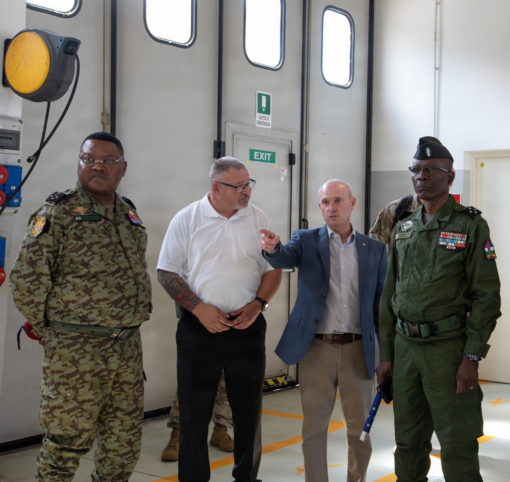Four individuals are standing inside what appears to be a military or industrial facility. Two men are in military uniforms with rank insignia and medals, one is in civilian business attire (blue blazer), and one is in a white polo shirt. The man in the blue blazer appears to be pointing and explaining something to the group.