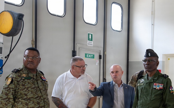 Four individuals are standing inside what appears to be a military or industrial facility. Two men are wearing military uniforms with insignia and medals, while two men are in civilian attire. One civilian in a blue blazer is gesturing and appears to be explaining or directing attention toward something.