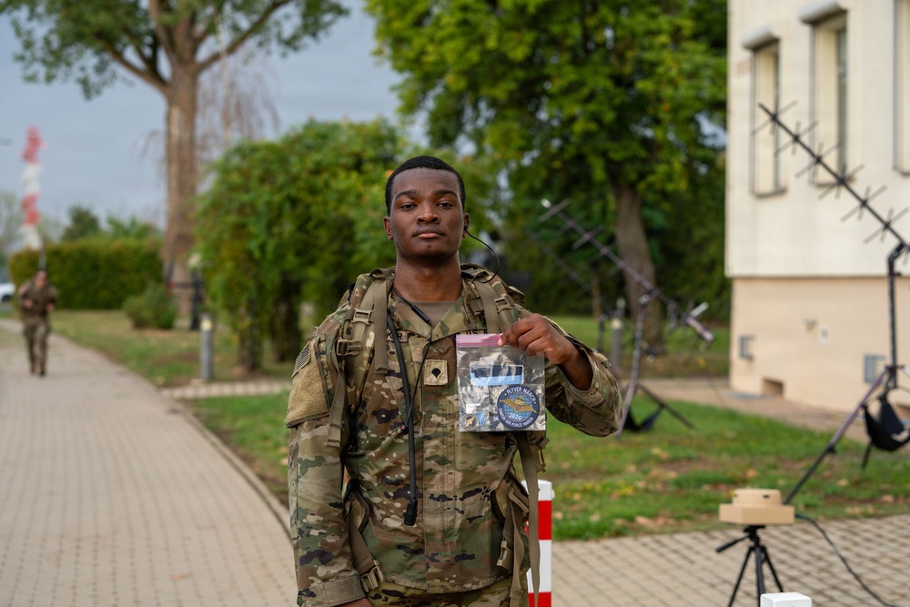 A U.S. military service member in OCP (Operational Camouflage Pattern) uniform is holding up a clear plastic bag containing what appears to be a military patch or coin, likely a commemorative item. The soldier is standing on a paved walkway on what appears to be a military installation, with trees, a building, and barbed wire visible in the background. Another service member can be seen walking in the background.