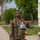 12th Combat Aviation Brigade hosts Danish Flyver March A U.S. military service member in OCP (Operational Camouflage Pattern) uniform is holding up a clear plastic bag containing what appears to be a military patch or coin, likely a commemorative item. The soldier is standing on a paved walkway on what appears to be a military installation, with trees, a building, and barbed wire visible in the background. Another service member can be seen walking in the background.