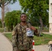 A U.S. military service member in OCP (Operational Camouflage Pattern) uniform is holding up a clear plastic bag containing what appears to be a military patch or coin, likely a commemorative item. The soldier is standing on a paved walkway on what appears to be a military installation, with trees, a building, and barbed wire visible in the background. Another service member can be seen walking in the background.
