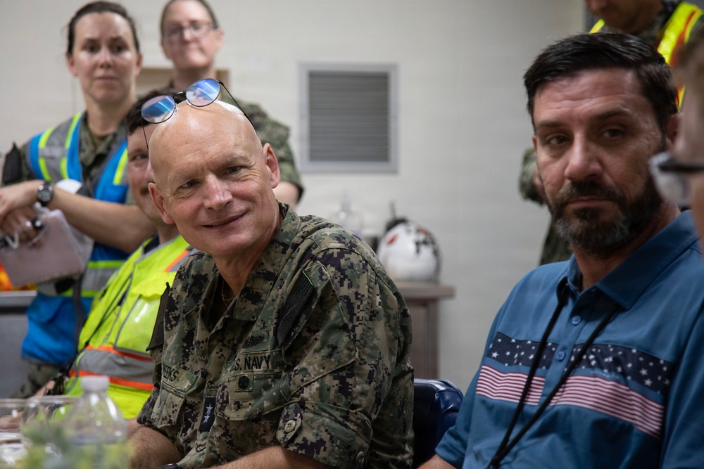 A group of individuals are gathered in what appears to be an indoor briefing or meeting room. A U.S. Navy officer in digital camouflage uniform is prominently seated in the foreground alongside a civilian man wearing a blue shirt with an American flag design. Several other personnel, including individuals in high-visibility vests and military uniforms, are visible in the background.
