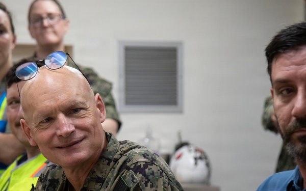A group of individuals are gathered in what appears to be an indoor briefing or meeting room. A U.S. Navy officer in digital camouflage uniform is prominently seated in the foreground alongside a civilian man wearing a blue shirt with an American flag design. Several other personnel, including individuals in high-visibility vests and military uniforms, are visible in the background.