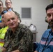 A group of individuals are gathered in what appears to be an indoor briefing or meeting room. A U.S. Navy officer in digital camouflage uniform is prominently seated in the foreground alongside a civilian man wearing a blue shirt with an American flag design. Several other personnel, including individuals in high-visibility vests and military uniforms, are visible in the background.