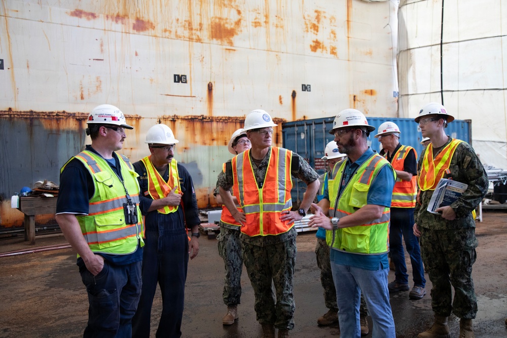 A group of approximately eight individuals are gathered in what appears to be a shipyard or dry dock facility, engaged in a briefing or discussion. The group includes personnel in U.S. Navy camouflage uniforms and civilians wearing high-visibility safety vests and white hard hats. A large rusted ship hull or structure dominates the background.