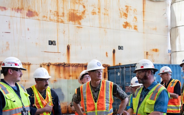 A group of approximately eight individuals are gathered in what appears to be a shipyard or dry dock facility, engaged in a briefing or discussion. The group includes personnel in U.S. Navy camouflage uniforms and civilians wearing high-visibility safety vests and white hard hats. A large rusted ship hull or structure dominates the background.