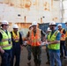 A group of approximately eight individuals are gathered in what appears to be a shipyard or dry dock facility, engaged in a briefing or discussion. The group includes personnel in U.S. Navy camouflage uniforms and civilians wearing high-visibility safety vests and white hard hats. A large rusted ship hull or structure dominates the background.