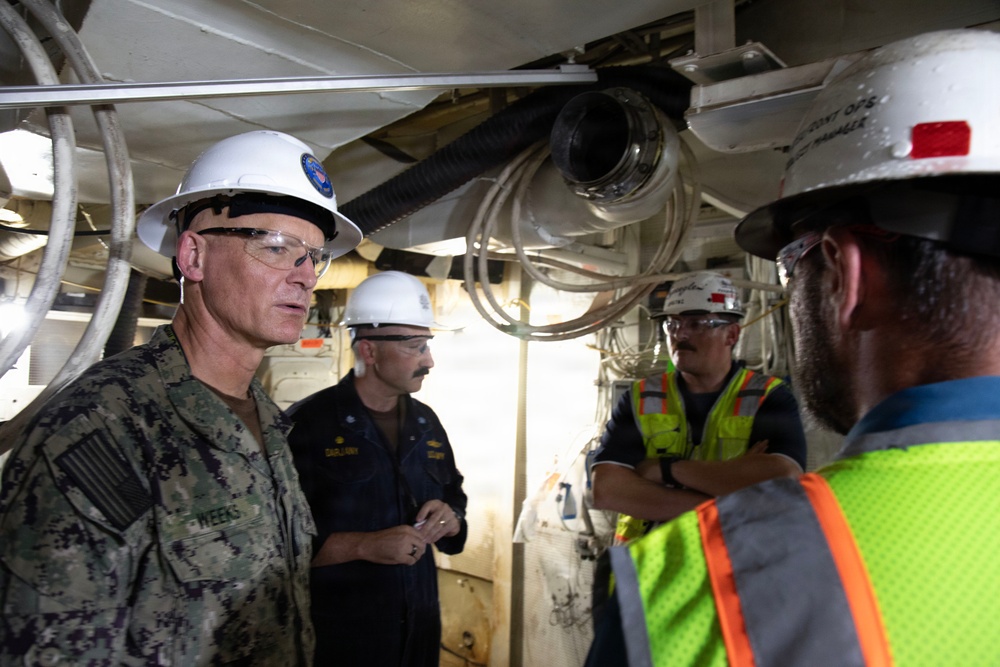 A group of military personnel and civilian workers are gathered in what appears to be an industrial or shipyard environment. A U.S. Navy officer in camouflage uniform with the name tag 'WEEKS' is speaking with others wearing white hard hats and safety gear. The setting suggests an inspection, briefing, or coordination meeting at a naval facility or shipyard.