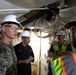 A group of military personnel and civilian workers are gathered in what appears to be an industrial or shipyard environment. A U.S. Navy officer in camouflage uniform with the name tag 'WEEKS' is speaking with others wearing white hard hats and safety gear. The setting suggests an inspection, briefing, or coordination meeting at a naval facility or shipyard.