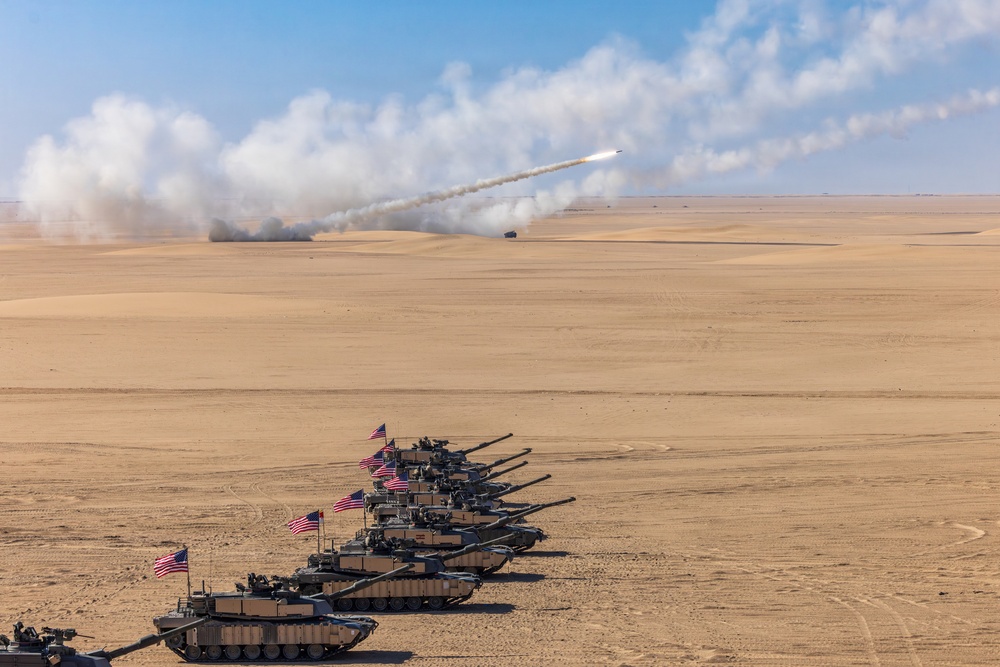 A formation of M1 Abrams main battle tanks bearing United States flags is lined up in a desert environment. In the background, a rocket or missile system (likely an M270 MLRS or HIMARS) is visibly launching a projectile, leaving a smoke trail across the sky. The scene appears to depict a military exercise or demonstration in an arid, open terrain.