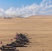 A formation of M1 Abrams main battle tanks bearing United States flags is lined up in a desert environment. In the background, a rocket or missile system (likely an M270 MLRS or HIMARS) is visibly launching a projectile, leaving a smoke trail across the sky. The scene appears to depict a military exercise or demonstration in an arid, open terrain.