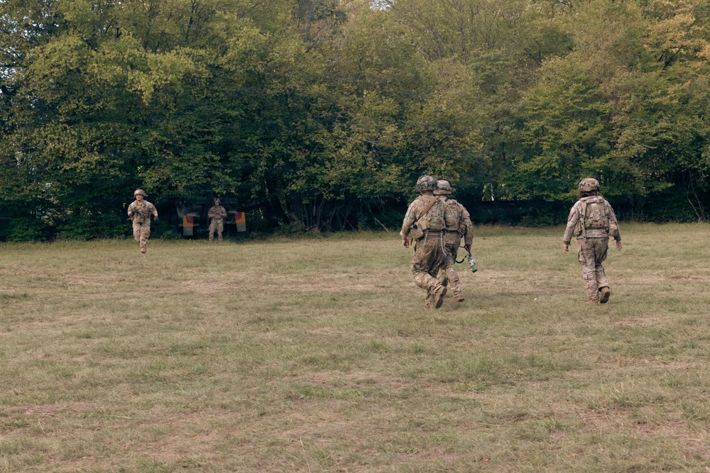The image shows multiple military personnel in camouflage uniforms and helmets moving across an open grassy field toward a tree line. Two soldiers in the foreground are advancing toward the wooded area, while two additional figures are visible in the background near what appears to be a colored marker or flag. The setting and equipment suggest a military training exercise.