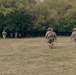 The image shows multiple military personnel in camouflage uniforms and helmets moving across an open grassy field toward a tree line. Two soldiers in the foreground are advancing toward the wooded area, while two additional figures are visible in the background near what appears to be a colored marker or flag. The setting and equipment suggest a military training exercise.