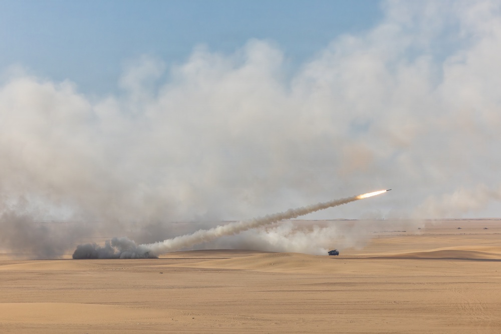 A rocket or missile has been launched from a desert environment, leaving a visible smoke trail across the sky. A military launch vehicle is visible on the ground near the center-right of the image. Large plumes of smoke and dust surround the launch area against a partly cloudy sky.