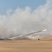 A rocket or missile has been launched from a desert environment, leaving a visible smoke trail across the sky. A military launch vehicle is visible on the ground near the center-right of the image. Large plumes of smoke and dust surround the launch area against a partly cloudy sky.
