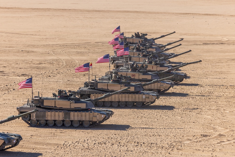 A formation of multiple M1 Abrams main battle tanks is arranged in a diagonal line in a desert environment. Each tank displays an American flag, and crew members are visible in the turret hatches. The formation appears to be a ceremonial or demonstration display, possibly during a military exercise or review.