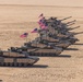 A formation of multiple M1 Abrams main battle tanks is arranged in a diagonal line in a desert environment. Each tank displays an American flag, and crew members are visible in the turret hatches. The formation appears to be a ceremonial or demonstration display, possibly during a military exercise or review.