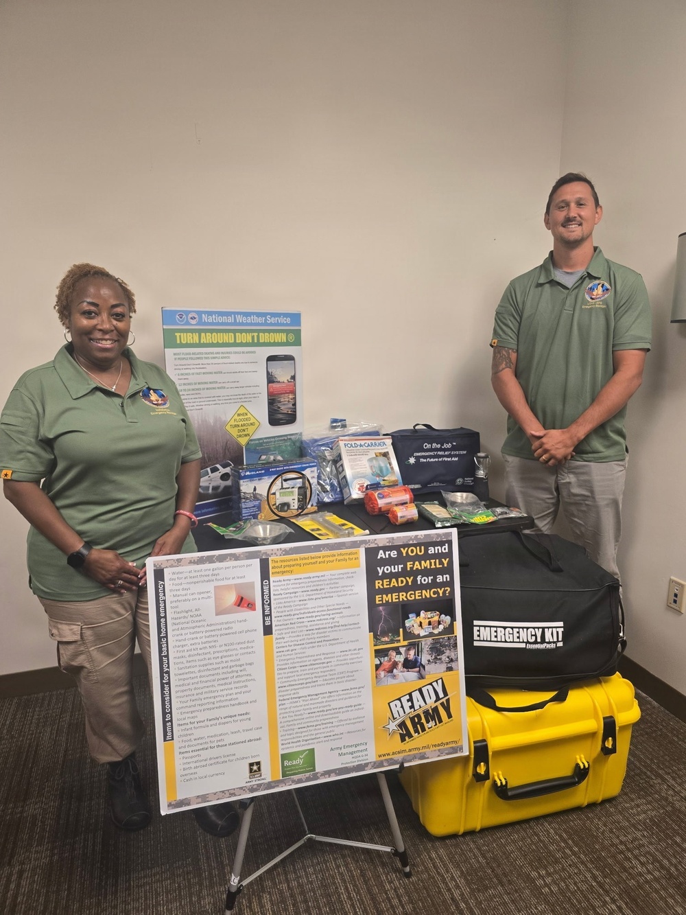 Two individuals in matching green polo shirts stand beside an emergency preparedness display table and signage. The display includes a 'Ready Army' poster, a National Weather Service 'Turn Around Don't Drown' poster, and various emergency kit items. A black and yellow hard-shell emergency kit case is prominently visible in the foreground.