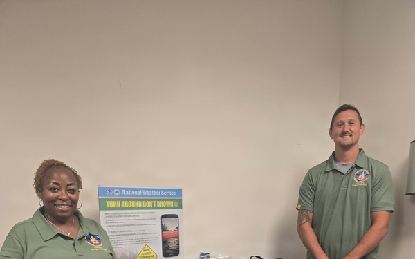 Two individuals in matching green polo shirts stand beside an emergency preparedness display table and signage. The display includes a 'Ready Army' poster, a National Weather Service 'Turn Around Don't Drown' poster, and various emergency kit items. A black and yellow hard-shell emergency kit case is prominently visible in the foreground.