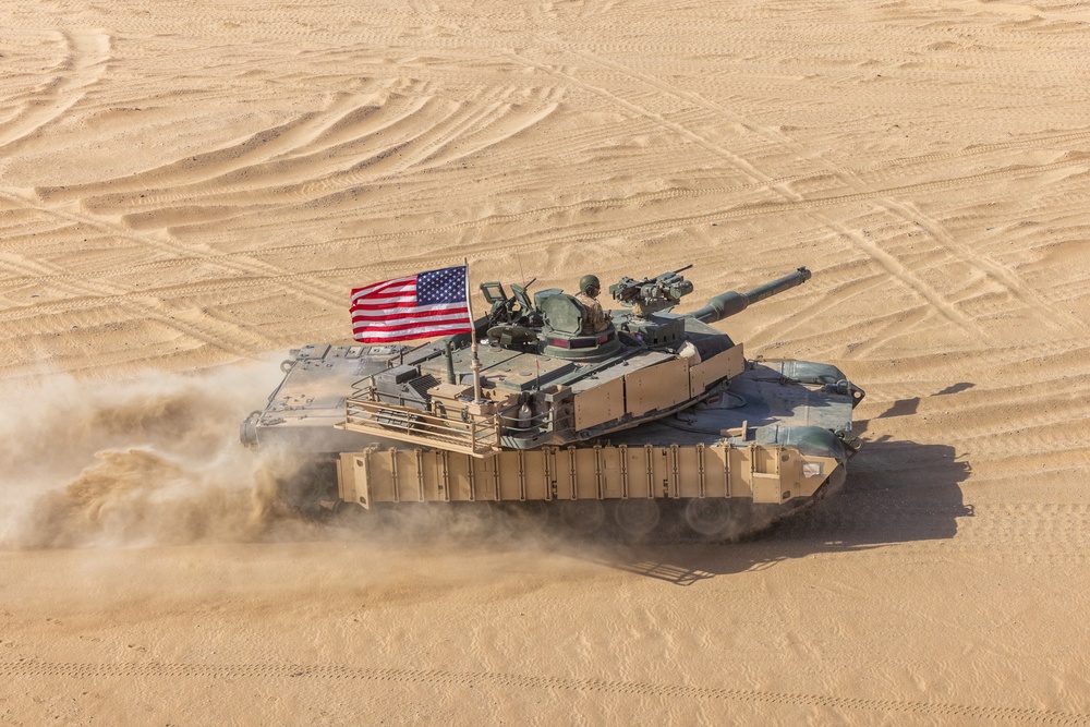 An aerial view of a U.S. Army M1 Abrams main battle tank moving at speed across a desert terrain, kicking up a dust cloud behind it. The tank is flying an American flag from its turret and appears to be in an active operational or training environment. The desert surface shows multiple vehicle track marks indicating prior activity in the area.
